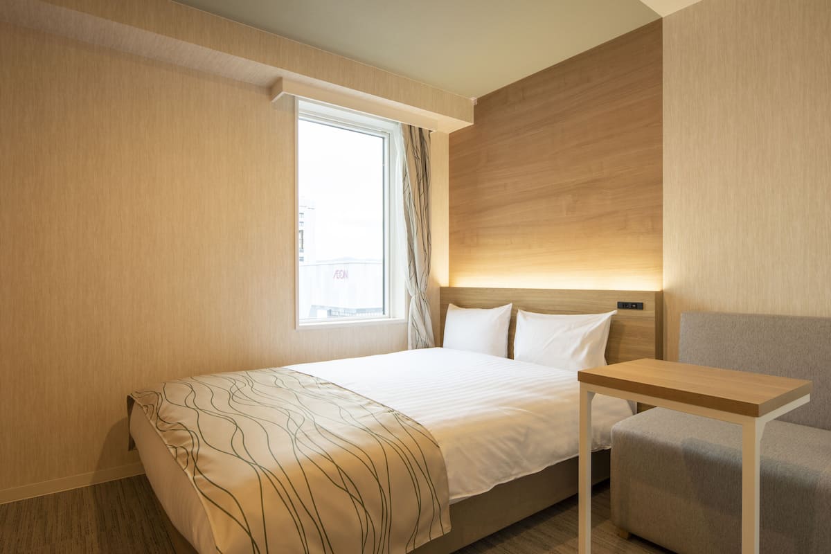 Une chambre d’hôtel avec un lit, une petite table, une chaise, une fenêtre avec des rideaux et un lampadaire fixé au mur.