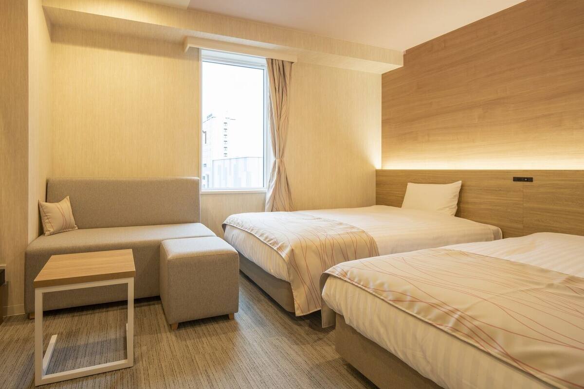Une chambre d’hôtel avec un canapé, deux lits simples, une petite table et une fenêtre avec des rideaux.
