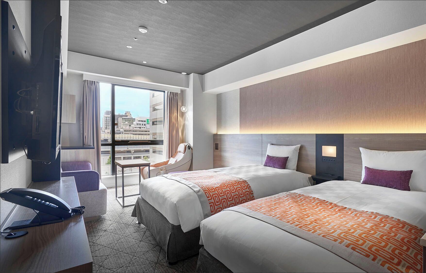 A hotel room with two beds, a desk, a chair, a phone, and a view of the city.
