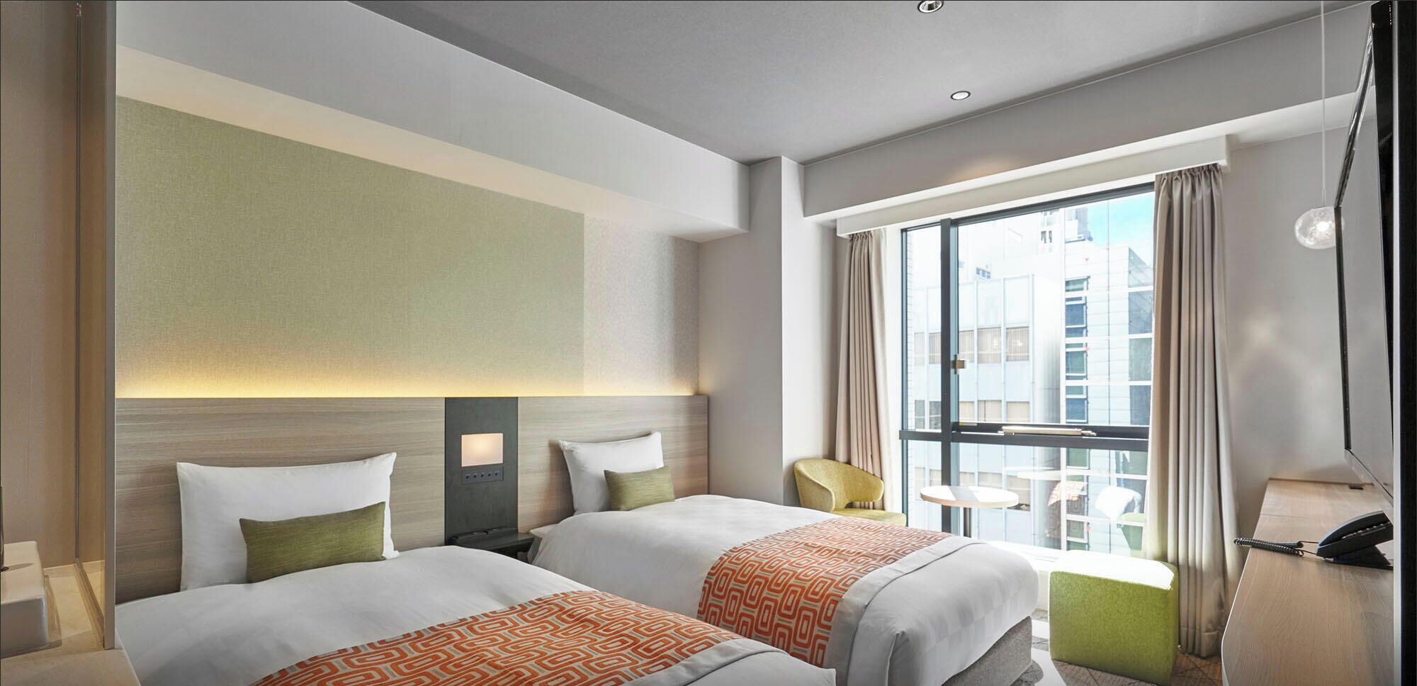 A hotel room with two beds, a balcony, a desk with a phone, and a view of the cityscape.