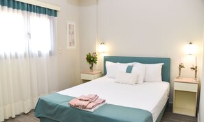 A neatly made bed with a teal bedspread and pink towels, two bedside tables with lamps, a window with sheer curtains, and a framed picture on the wall. - Aurora Luxury Retreat - Beachfront & Private Pool (Messini)