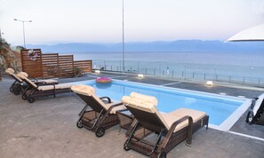 A poolside area with lounge chairs and a floating ring, overlooking a body of water with distant mountains. - Aurora Luxury Retreat - Beachfront & Private Pool (Messini)