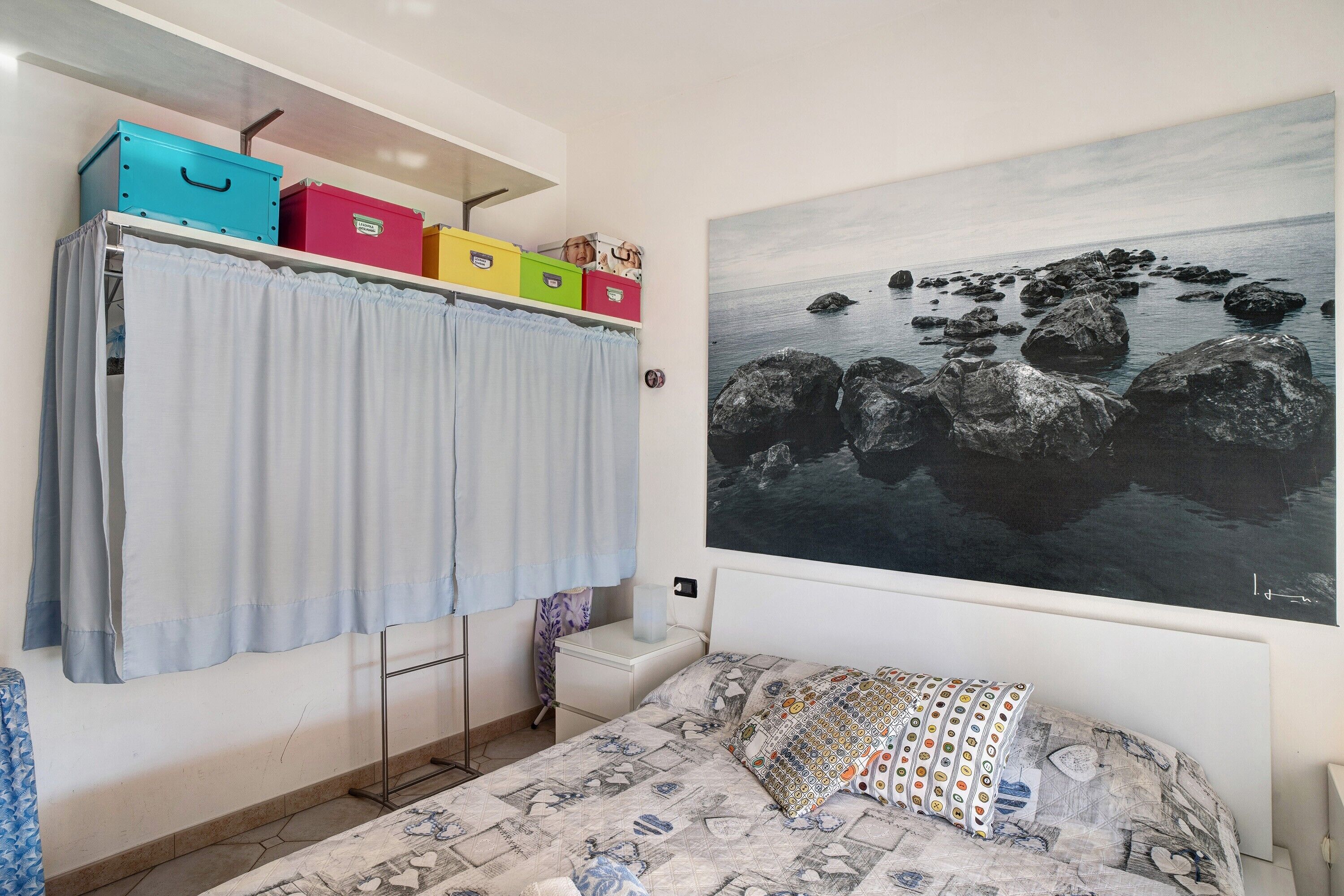 A bedroom with a bed, a wall-mounted shelf with storage boxes, a curtain, and a large landscape painting.