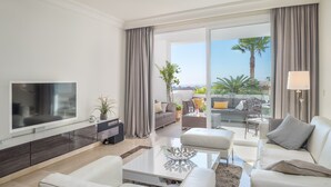 A modern living room with a flat-screen TV, a sofa, a coffee table, and a balcony with a view of palm trees. - Apartment "Seaview Flat Benahavis-Marbella" with Sea View, Pool, Wi-Fi, Terrace & Garden (Benahavis)