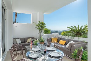 Un espace extérieur lumineux, aménagé pour les repas, avec une table en verre, des chaises en métal et un canapé avec des coussins.