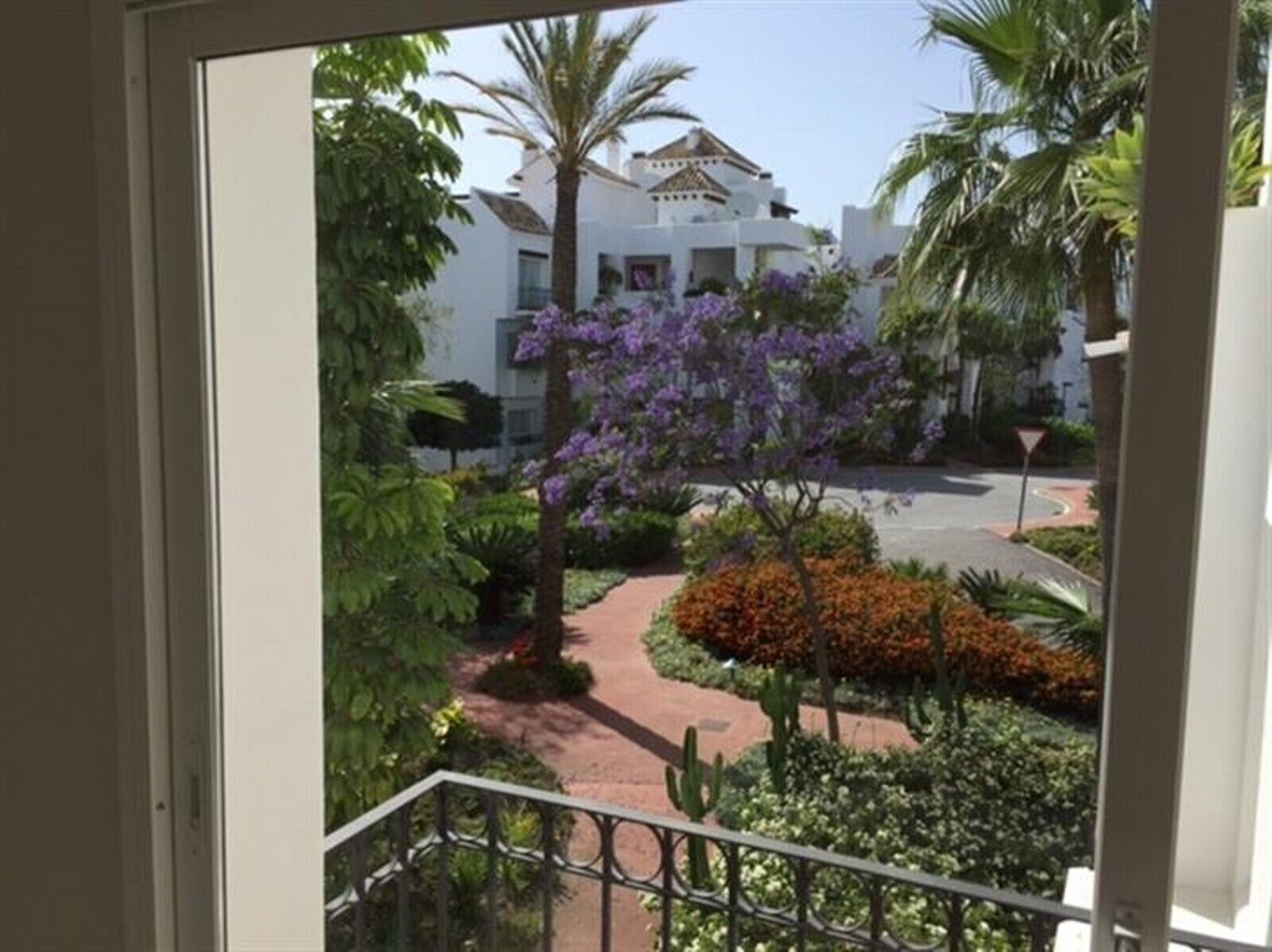 A balcony with a view of a garden, palm trees, and a building with white walls and a brown roof.