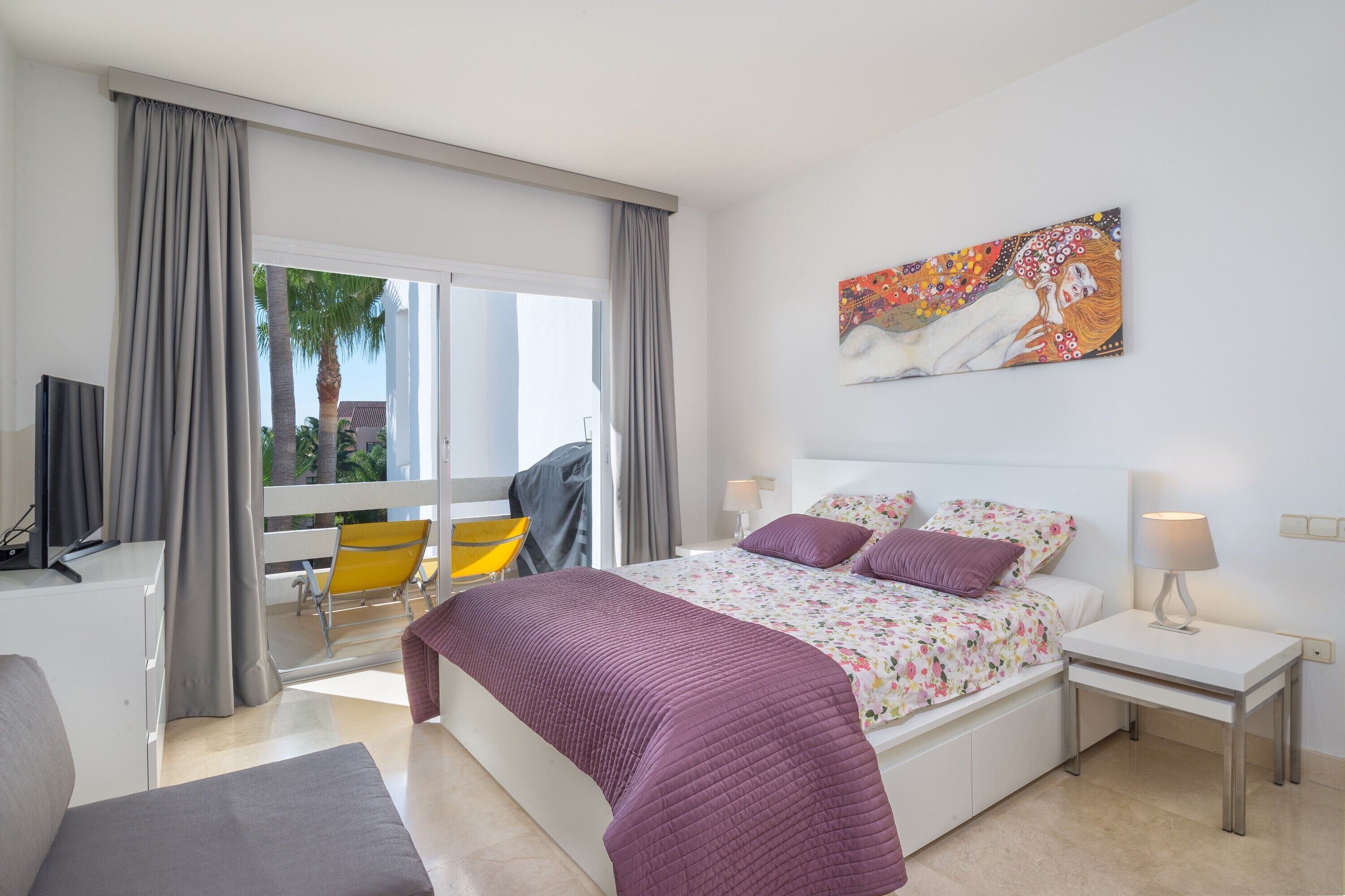 Een slaapkamer met een bed, nachtkastjes, een stoel, een televisie en een balkon met een stoel en gordijnen.