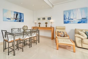 Un coin repas avec une table à plateau de verre et des chaises, une table d’appoint en bois avec des lampes, et deux tableaux représentant des voiliers accrochés aux murs.