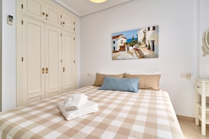 A bedroom with a checkered bedspread, a painting of a street scene, a wardrobe, and a bedside table.