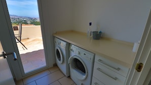 A laundry area with two washing machines, a small table with a water bottle and a basket, and a view of a cityscape through an open door. - 'Luxurious villa in the sun' with Sea & Mountain View, Pool, A/C & Wi-Fi (Mijas Costa)