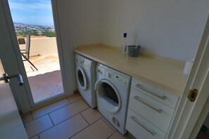 Zona de lavandería con dos lavadoras, una mesita con una botella de agua y una cesta, y vista a un paisaje urbano a través de una puerta abierta.