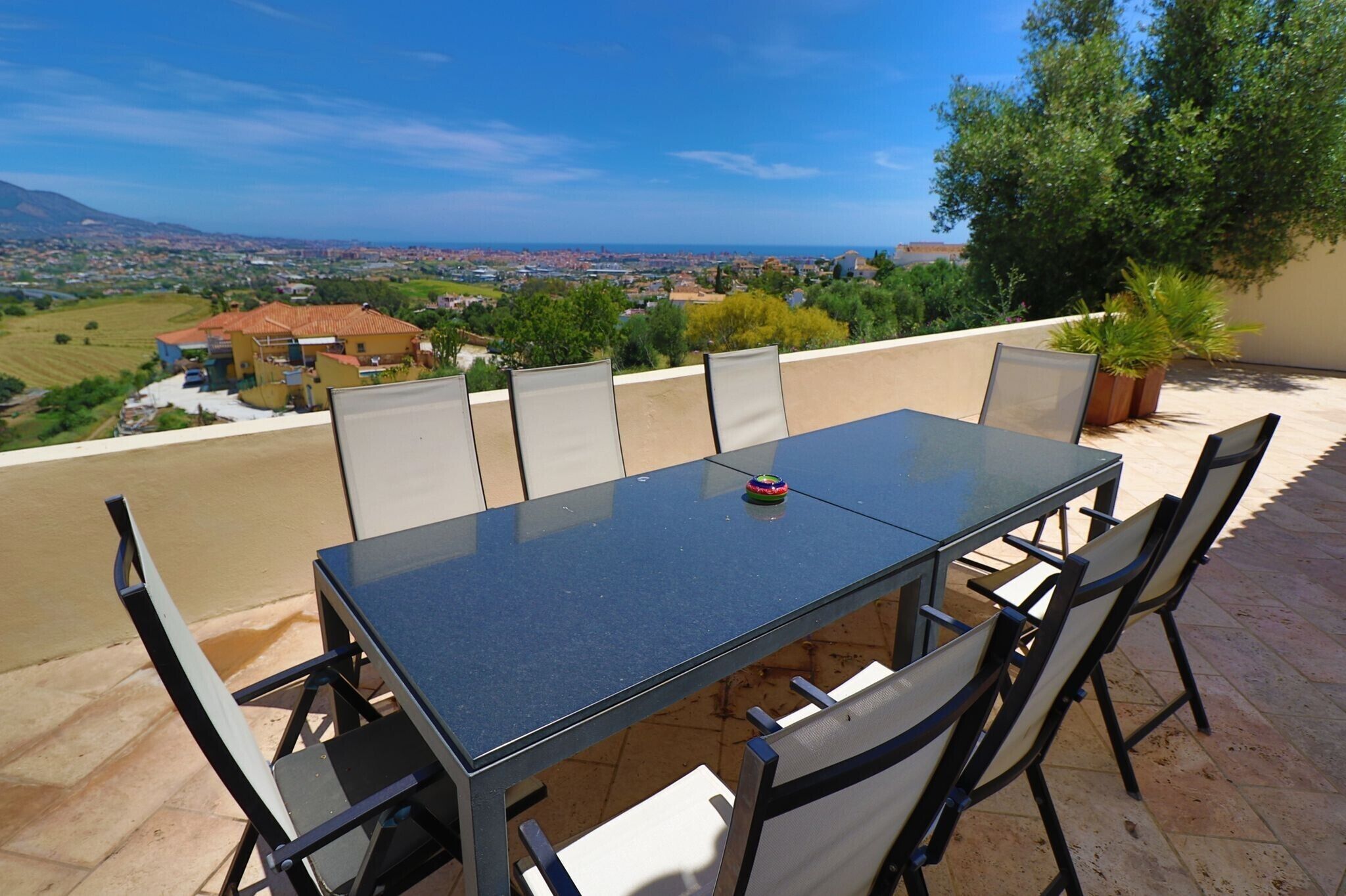 An outdoor dining area with a table and chairs, a potted plant, and a view of a town and mountains.