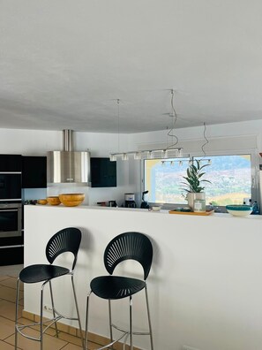 Uma cozinha moderna com uma ilha grande, dois bancos altos pretos, uma coifa de aço inoxidável e uma janela com vista para a paisagem.