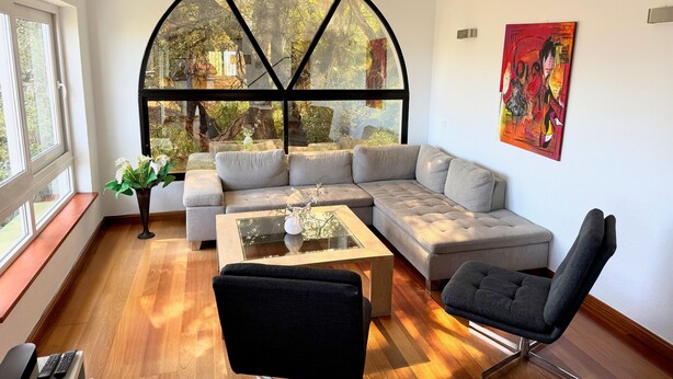 A modern living room with a sectional sofa, a glass coffee table, a black armchair, and a large arched window offering a view of trees and a building. - 'Nice and Spacius Villa near Fuengi' with Sea & Mountain View, Pool, NO under 40 (Mijas)