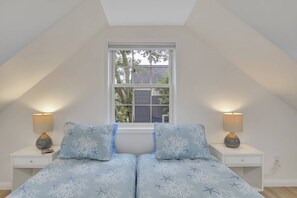 Une chambre avec un plafond en pente, une fenêtre donnant sur les arbres, deux tables de chevet avec des lampes et un lit recouvert d’une couvre-lit à motifs bleus et blancs.