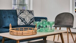 Een woonkamer met een blauwe bank, een kussen met dessin, een houten bijzettafeltje en een stoel.