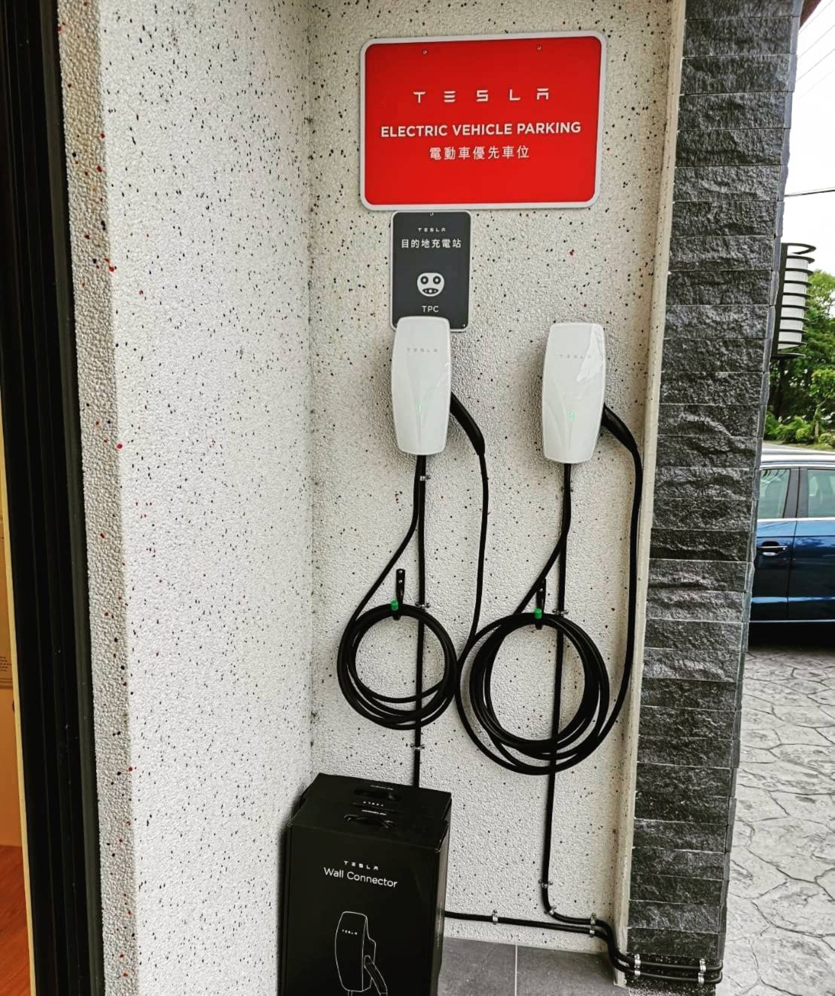 A Tesla electric vehicle charging station with a sign indicating it is for Tesla vehicles, a TPC charging icon, and a wall connector box.
