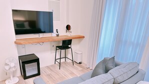 A modern hotel room with a grey sofa, a wooden desk with a flat-screen TV, a small black chair, a white trash bin, and a standing fan. - Gardenstay (Luodong)