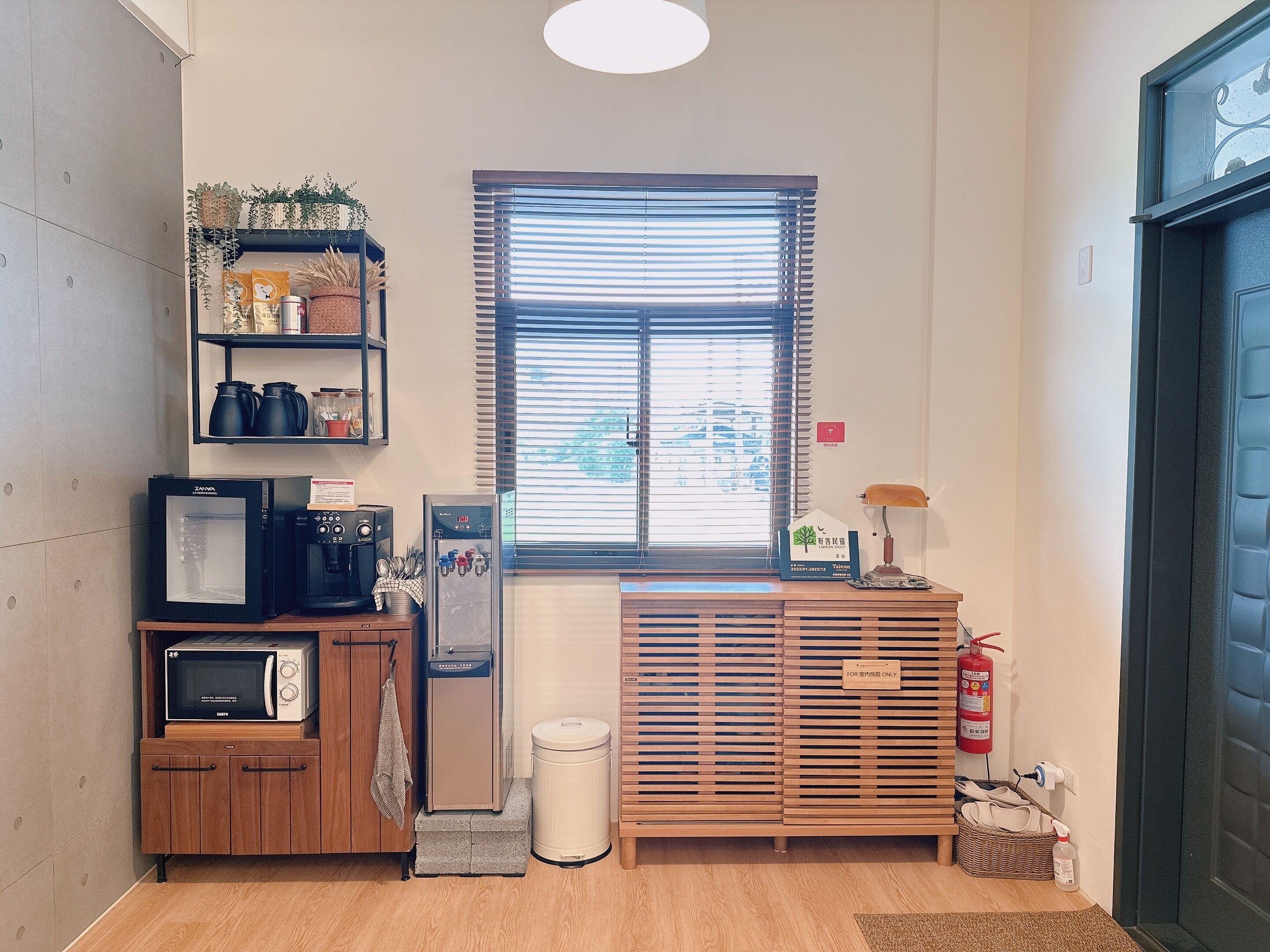 A compact kitchenette with a microwave, toaster, and coffee maker, a small trash bin, a window with blinds, and a fire extinguisher.