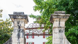 A stone gate with a wrought iron gate, leading to a white building with red shutters and a small garden. - The noble house of Luroa built in 1505. (Cambo-les-Bains)