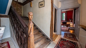 A wooden staircase with a carved figure, a framed picture on the wall, and a red door leading to a dining area. - The noble house of Luroa built in 1505. (Cambo-les-Bains)