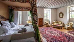 A bedroom with a four-poster bed, a large window, a wooden dresser, a floral armchair, and a patterned rug. - The noble house of Luroa built in 1505. (Cambo-les-Bains)