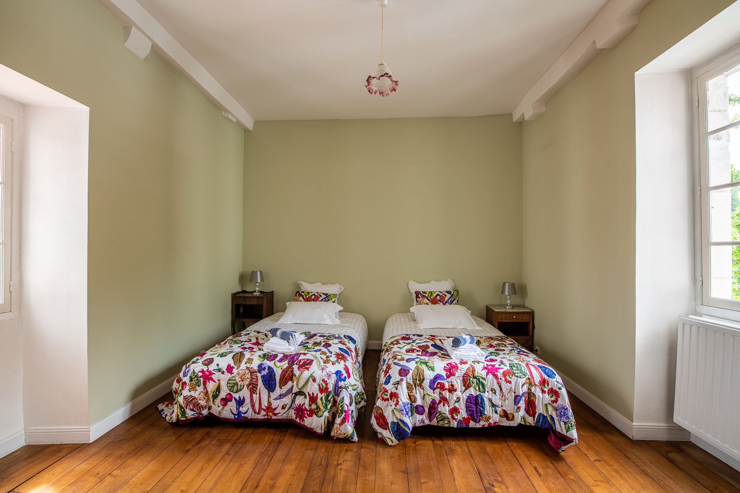 Quarto com duas camas, cada uma com roupa de cama floral, piso de madeira e uma pequena mesa de cabeceira com abajur.