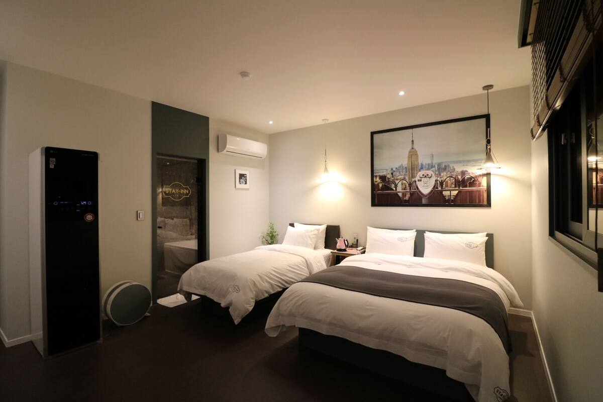 A hotel room with two beds, a wall-mounted air conditioner, a door with a 'Stay-Inn' sign, a framed cityscape picture, and a window with blinds.