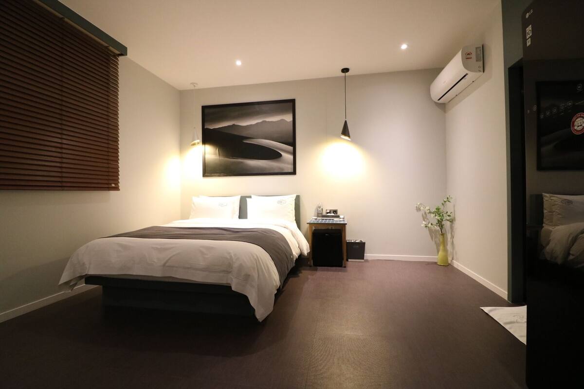 A bedroom with a bed, bedside table, lamp, air conditioning unit, and a painting on the wall.