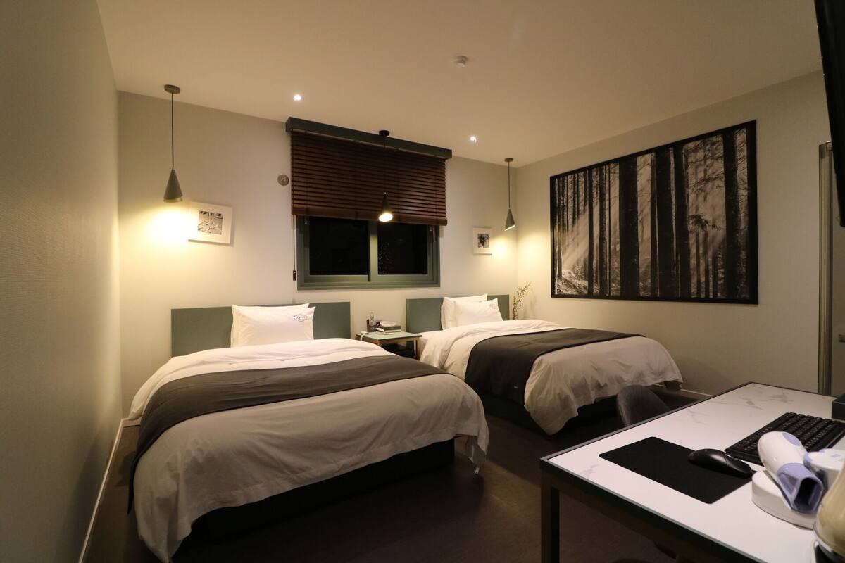 A hotel room with two beds, a desk with a computer, and a large forest-themed painting on the wall.