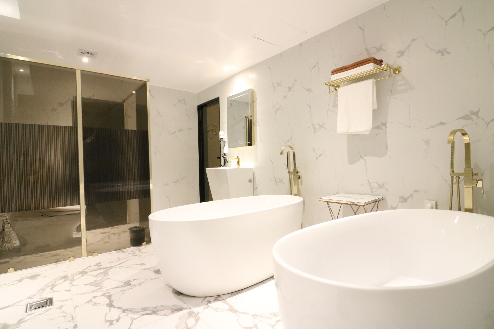 Um banheiro moderno com duas banheiras grandes e brancas, chuveiro com box de vidro e piso de mármore.
