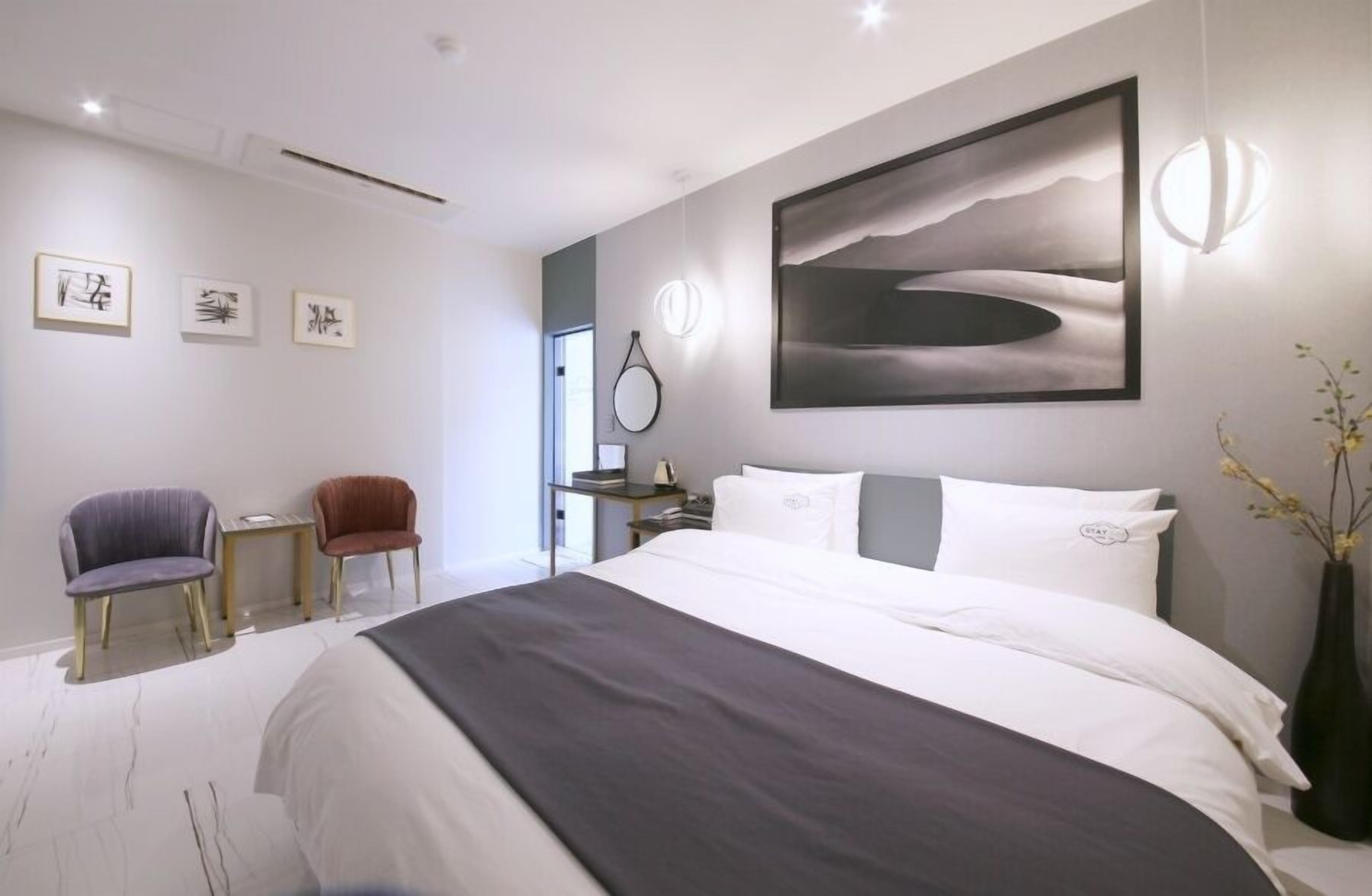 A modern hotel room with a large bed, a desk, two chairs, a vase with flowers, and artwork on the walls.