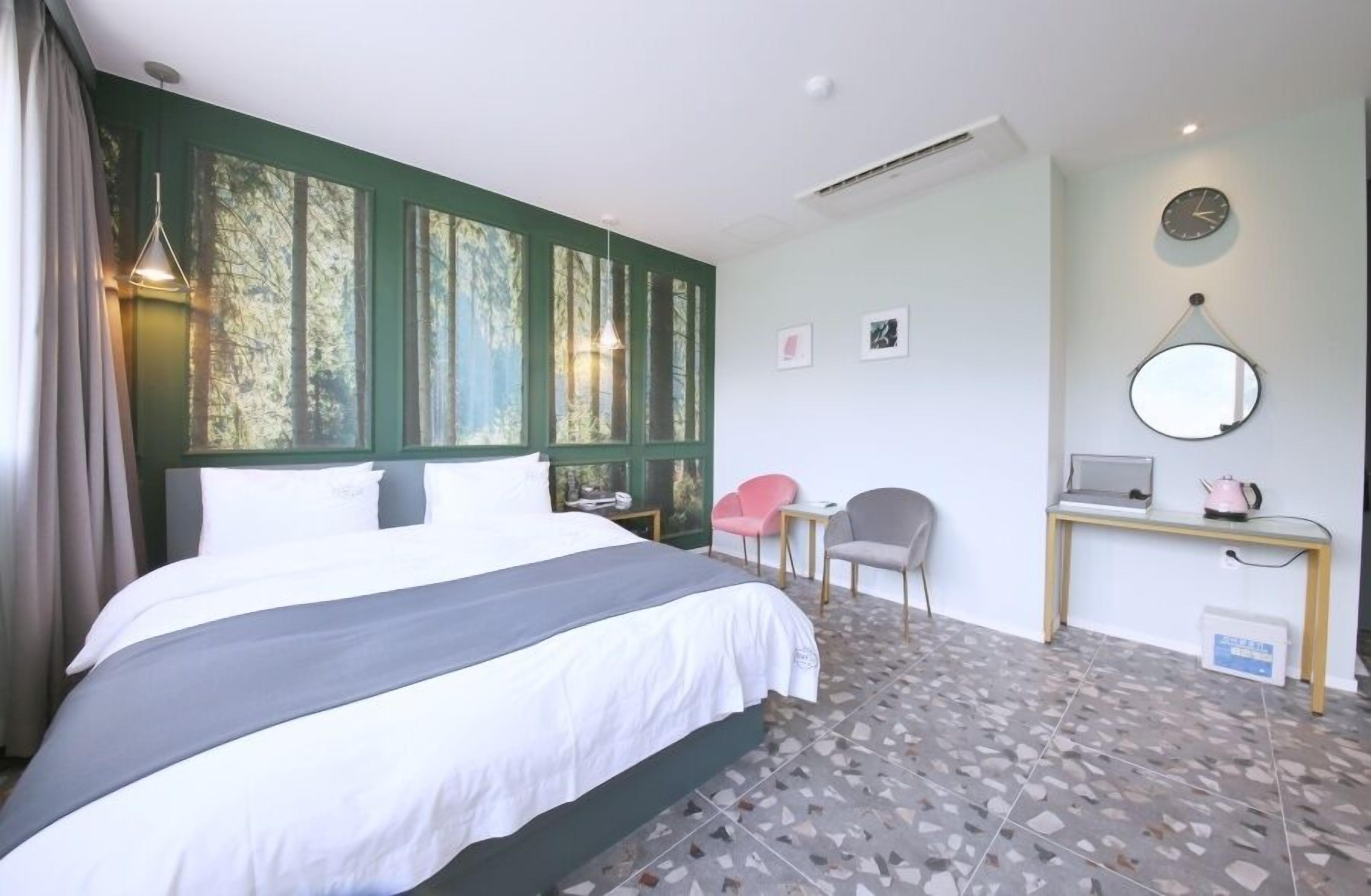 Ein Hotelzimmer mit einem großen Bett, einem Schreibtisch mit Computer, einem Sessel und einem kleinen Tisch mit Wasserkocher.