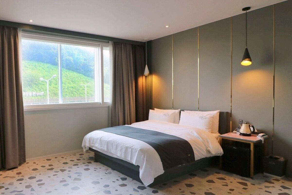 A modern hotel room with a large bed, a bedside table with a lamp, and a view of a green landscape through the window.