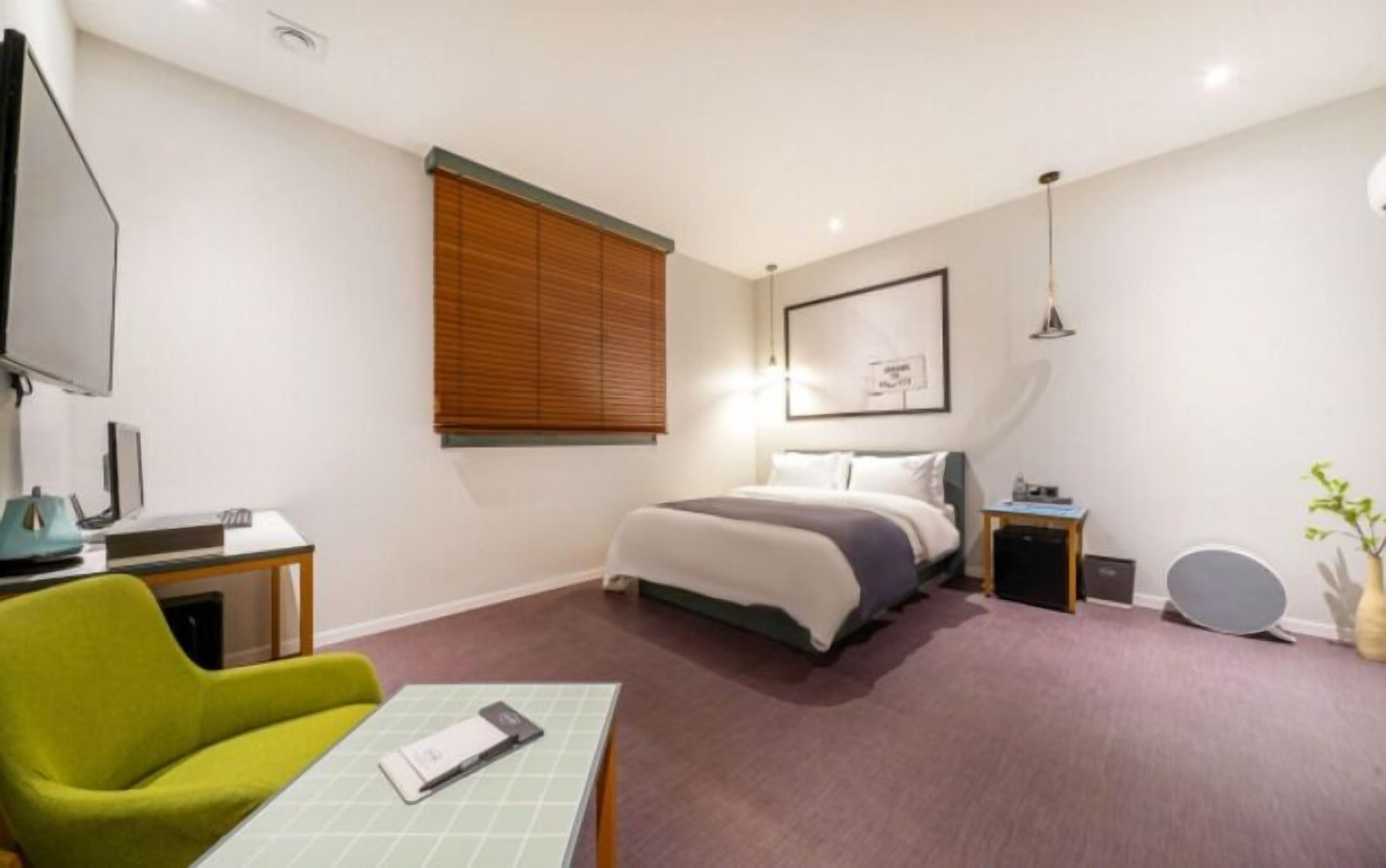 Habitación de hotel con cama, escritorio con computadora, silla, mesita de noche y televisor instalado en la pared.