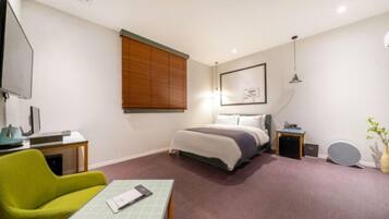 Habitación de hotel con cama, escritorio con computadora, silla, mesita de noche y televisor instalado en la pared.
