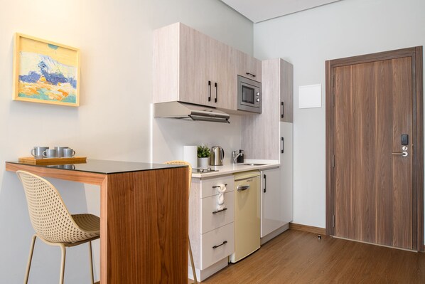 Una kitchenette compacta con microondas, estufa y pileta. Hay una silla de madera y una mesita con tazas. Se ve una puerta a la derecha.