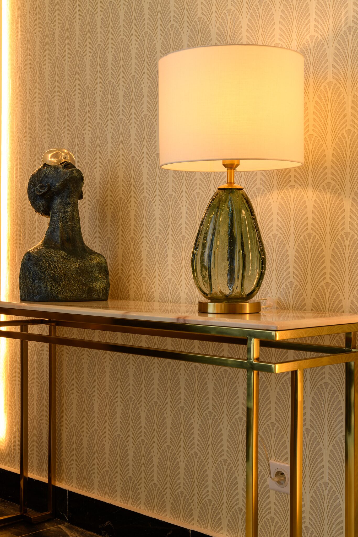 Une pièce avec un papier peint à motifs, une table en verre avec un cadre en laiton et une lampe avec un abat-jour blanc.