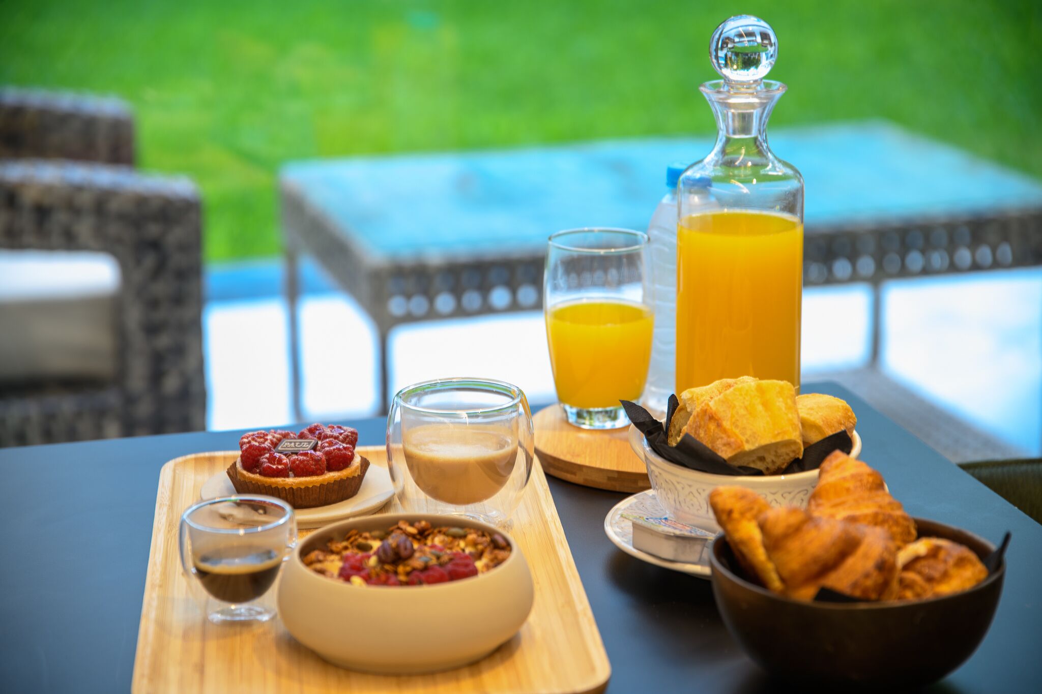 Ein Frühstücksbrett mit einer Schüssel Müsli, einem Glas Saft, einer Tasse Kaffee, einem Gebäckstück und einer Orangensaftflasche auf einem Tisch.