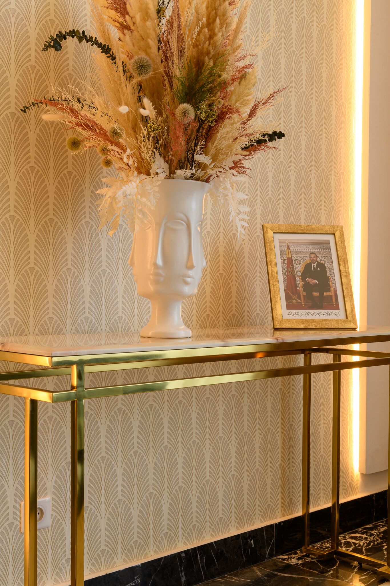 A white vase with dried plants on a glass table against a wallpapered wall.