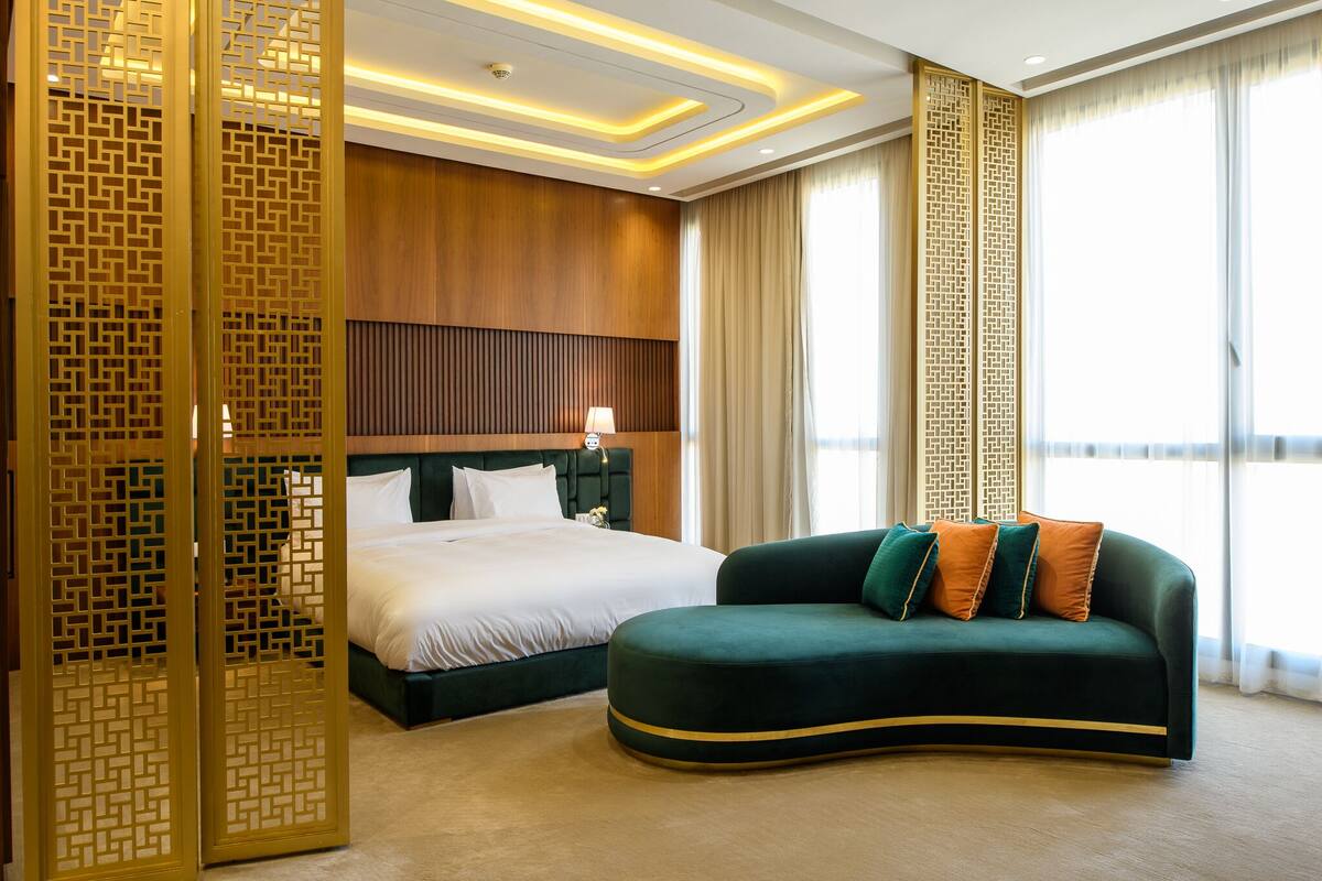 Quarto de hotel com cama, sofá verde, divisória decorativa e uma grande janela com cortinas.