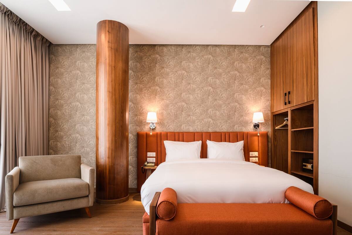 Quarto de hotel com uma cama grande, um guarda-roupa de madeira, uma cadeira e uma mesa de cabeceira com abajur.