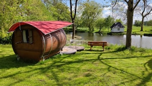A wooden barrel house with a red roof, a small deck with chairs and a table, a bench, and a pond in the background.