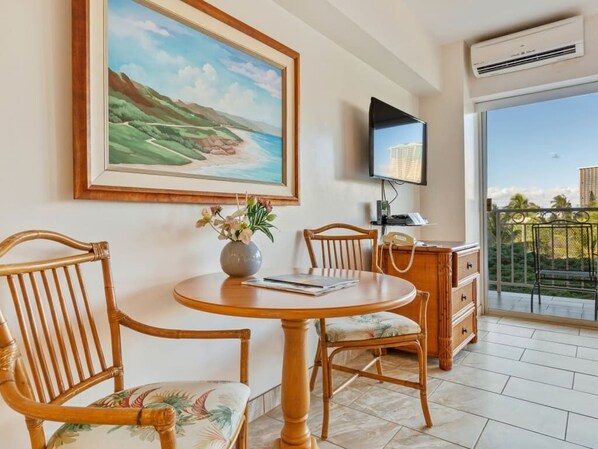 A room with a round wooden table, two chairs, a vase with flowers, a framed painting, a flat-screen TV, a desk with a telephone, and a view of the outdoors through a glass door.