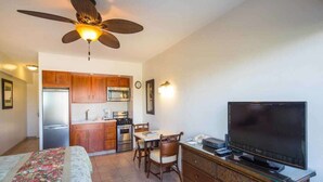 A compact room with a bed, a television, a dining table with chairs, a kitchen area with cabinets and appliances, and a ceiling fan.