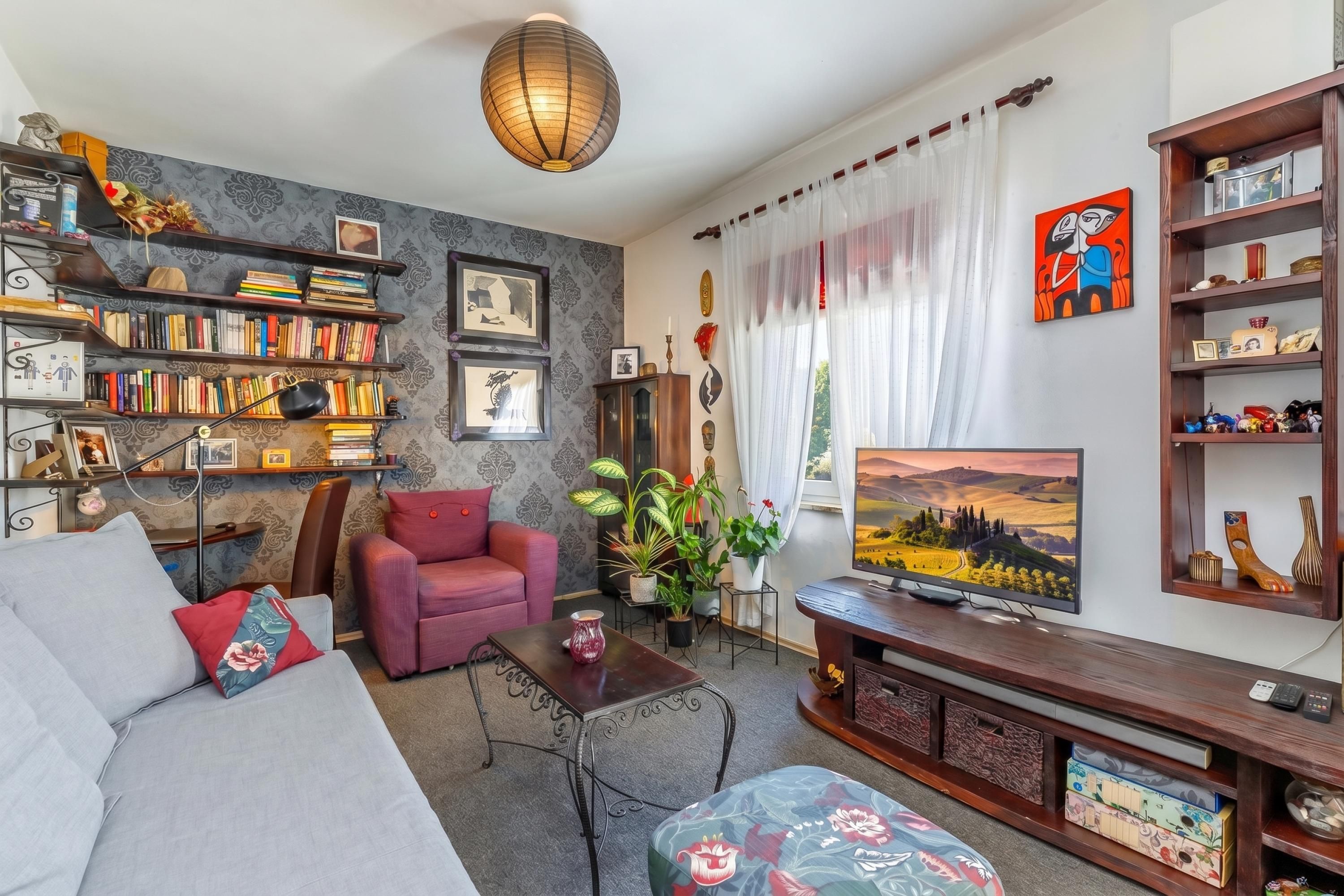 Una sala de estar con un sofá, un sillón rojo, una mesa de centro, un televisor y una estantería llena de libros y objetos decorativos.