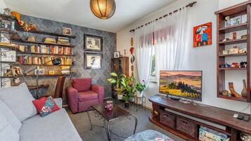 Una sala de estar con un sofá, un sillón rojo, una mesa de centro, un televisor y una estantería llena de libros y objetos decorativos.