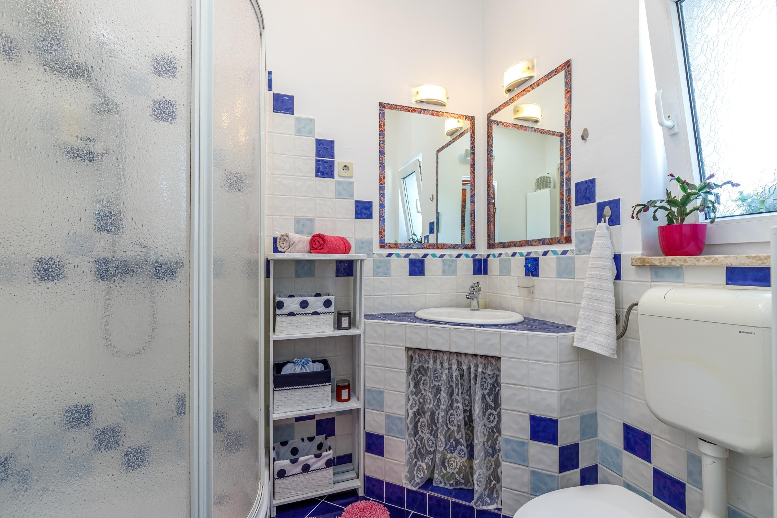 Un baño con inodoro blanco, lavamanos, dos espejos con marcos decorativos, estante con toallas y cestas, y ventana con vistas.