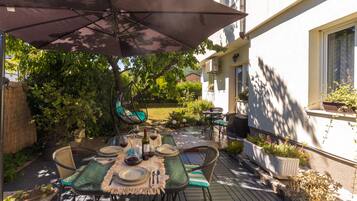 An outdoor patio with a table set for dining, surrounded by greenery and a house.