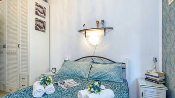 A neatly made bed with patterned bedding, a bedside table with a lamp, a nightstand with books, and a wall-mounted shelf with decorative items.